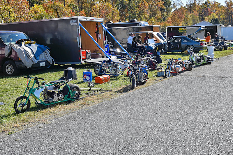 A row of small custom bikes parked alongside a gravel road, with trailers and vehicles in the background. The scene includes people socializing and working on the bikes amidst autumn trees.