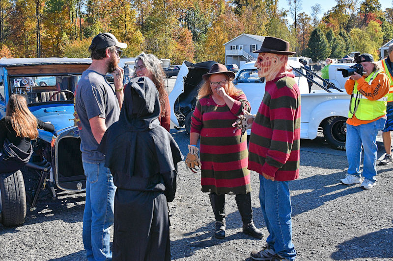 A group of people dressed in Halloween costumes at a car show, including individuals portraying iconic horror characters. One person in a dark cloak stands among others in striped sweaters, resembling a well-known slasher character, while an onlooker takes photos.