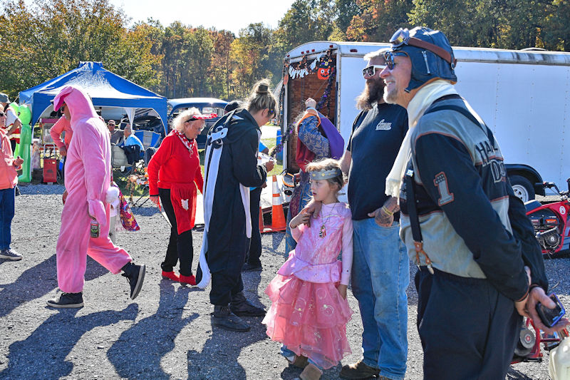 A colorful outdoor gathering featuring people in various costumes, including a pink outfit, a princess dress, and a pilot uniform, with tents and decorations in the background.