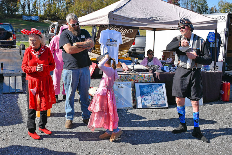 A colorful scene at an outdoor market with four individuals interacting. On the left, an elderly woman in a red dress and polka dot headband. A middle-aged man with a beard and black shirt stands beside her, while a young girl in a pink dress plays nearby. To the right, a man in a unique outfit with goggles shows a prop. A vendor tent displays various items for sale in the background.