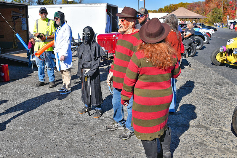A group of people in costumes at an outdoor event, featuring two individuals in striped sweaters and hats resembling a horror character, a child in a dark cloak with a mask, and others in casual attire. There are vintage cars and a trailer in the background, surrounded by fall foliage.