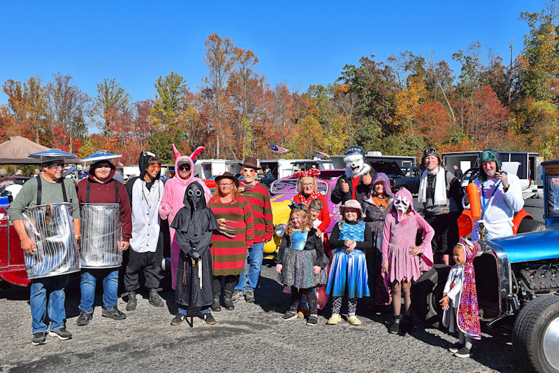 Group of people dressed in various costumes, posing together in front of colorful vintage cars with autumn trees in the background.