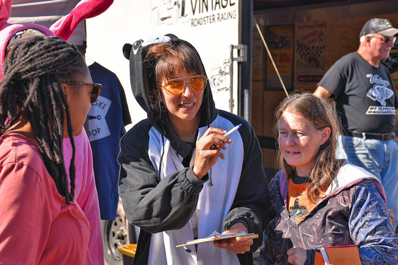 A woman dressed in a panda onesie holds a clipboard and pen, engaging in conversation with another woman, while a person wearing bunny ears stands nearby. The background features a vintage racing truck.
