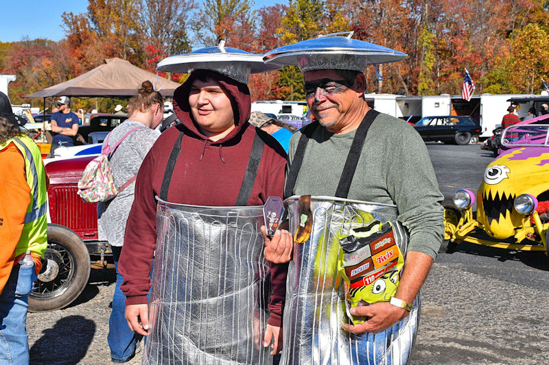 Two men dressed in metallic trash can costumes with lids on their heads, smiling and posing at an outdoor event, surrounded by colorful cars and autumn foliage.