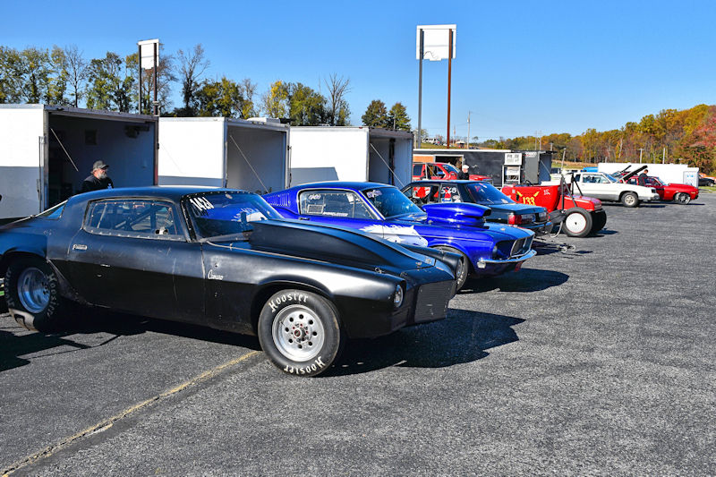 A row of classic and modified cars parked on a racetrack, with trailers in the background and a clear blue sky.