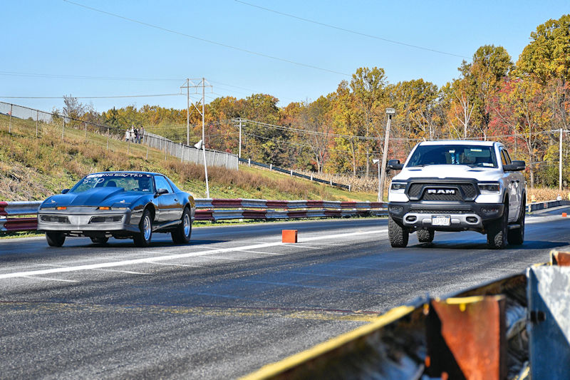 A silver car and a white truck racing on a drag strip, with autumn trees in the background.