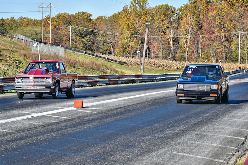 Two pickup trucks racing down a drag strip on a clear day, with trees displaying autumn colors in the background.