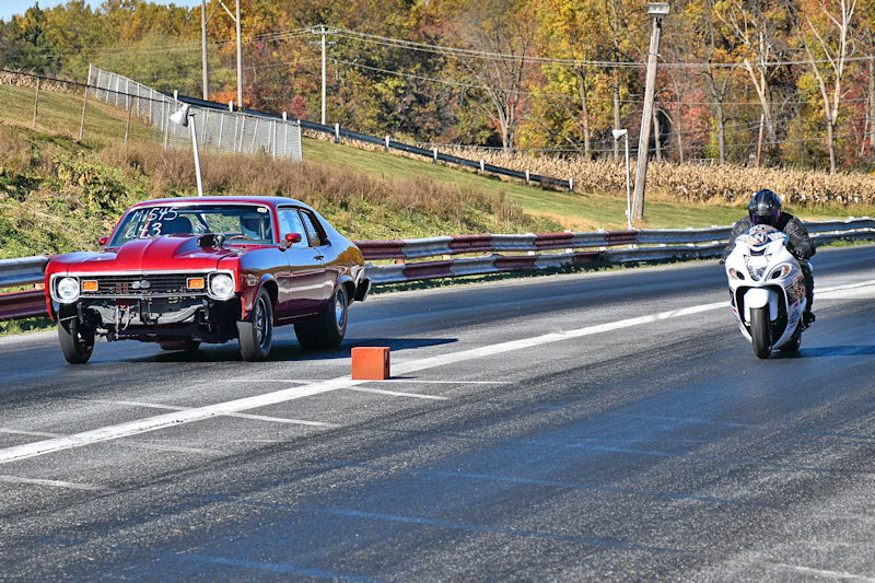 A classic muscle car and a sport motorcycle racing on a drag strip, with autumn foliage in the background.