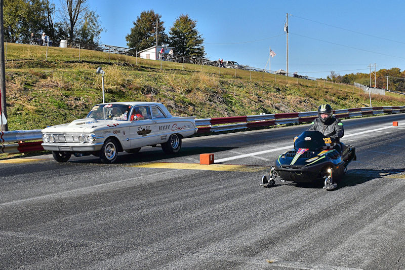 A vintage race car and a snowmobile lined up on a drag racing track, with spectators in the background and clear blue skies.