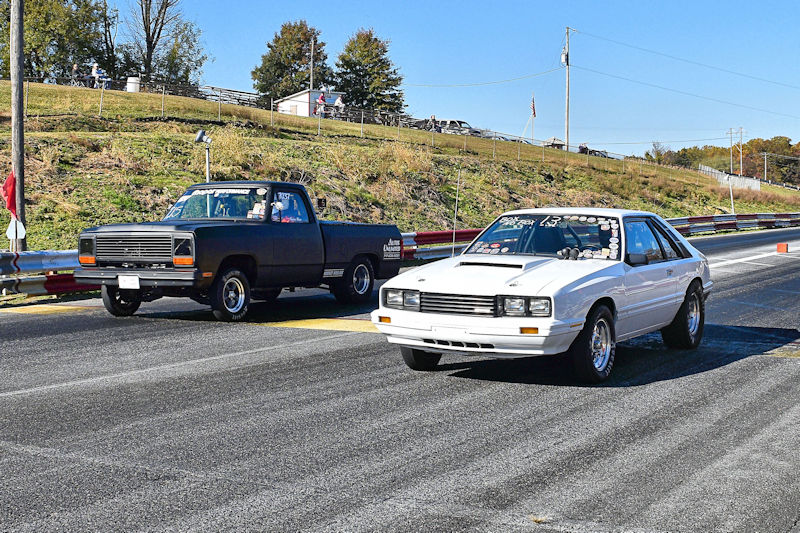 A black pickup truck and a white sports car lined up at a drag racing track, ready to race on a clear day.