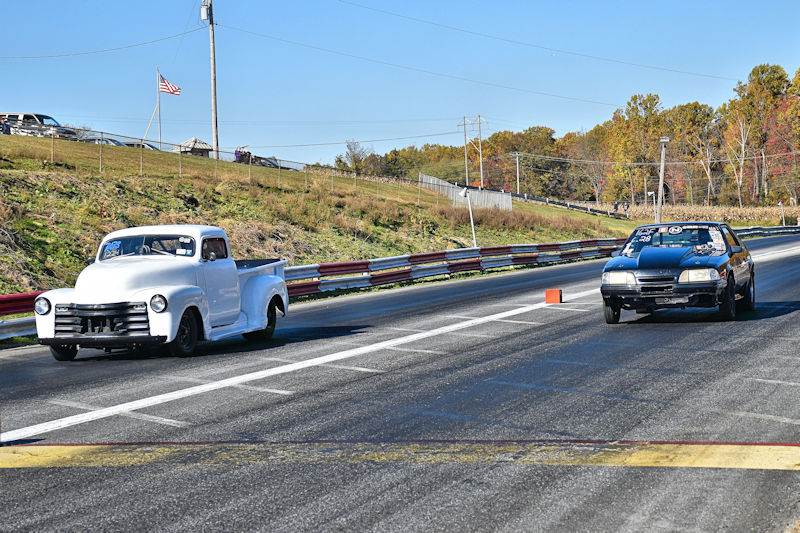 A white vintage pickup truck races alongside a black muscle car on a drag strip, with trees and an American flag in the background.