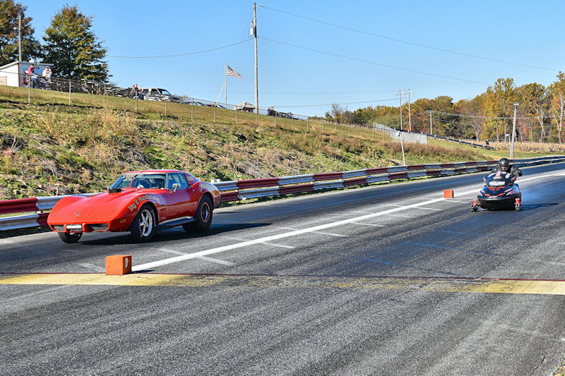 A red sports car and a racing kart compete on a drag strip with spectators in the background.