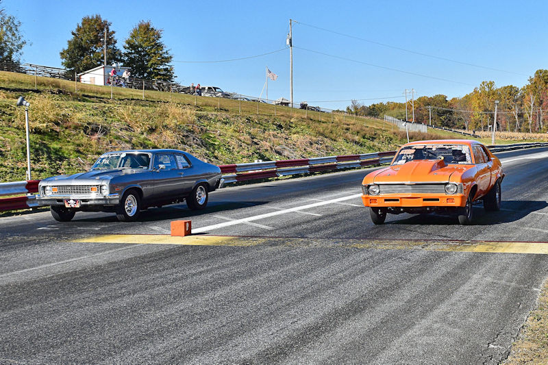 Two vintage muscle cars, one gray and one orange, are lined up at the starting line of a drag racing track on a sunny day.