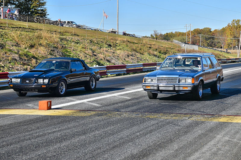 Two cars racing on a drag strip, with a black sports car on the left and a silver station wagon on the right.