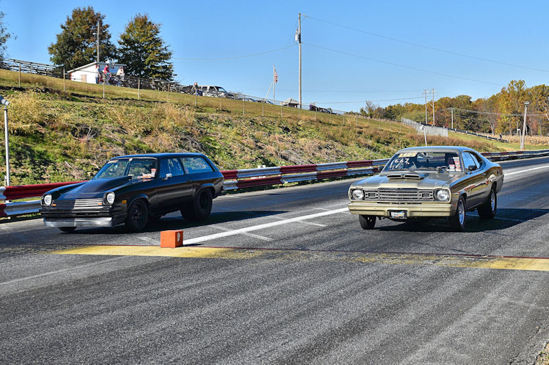 Two cars racing down a drag strip on a sunny day, with a wooded area in the background.