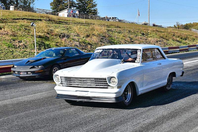 Two classic cars racing on a drag strip, a white car in the foreground and a black car in the background.