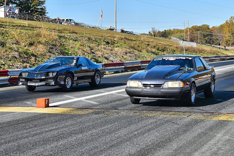 Two race cars competing on a drag strip, with one car in the left lane and another in the right lane. The background features a grassy area and a few spectators.