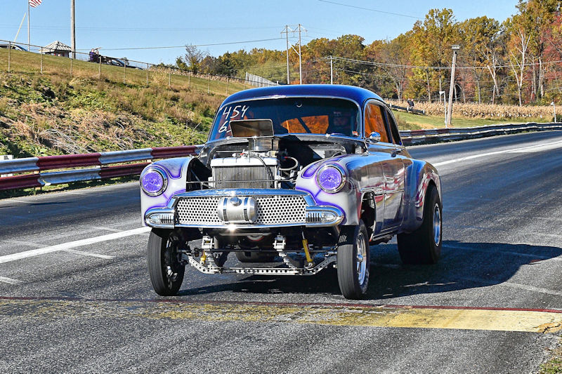 A vintage race car with a purple and silver exterior, featuring an exposed engine and large front tires, on a drag racing track.