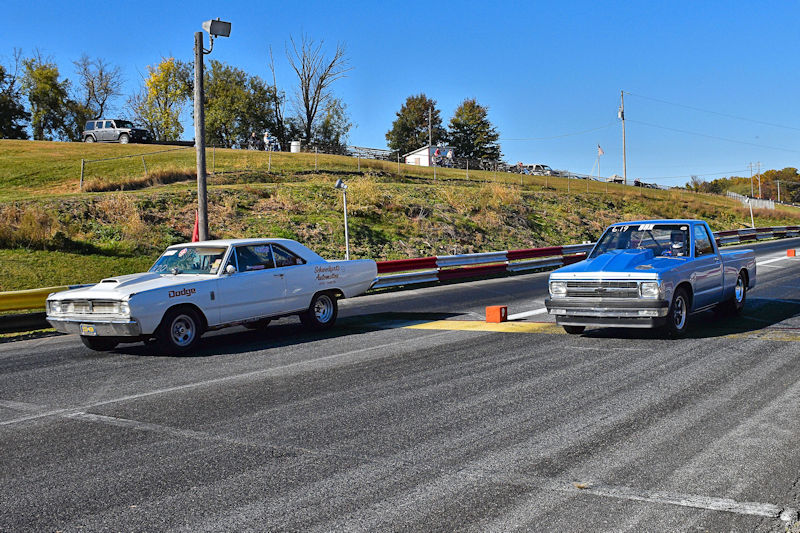 Two cars staged for a drag race on a racetrack, featuring a white Dodge and a blue pickup truck, with trees and a grassy area in the background.