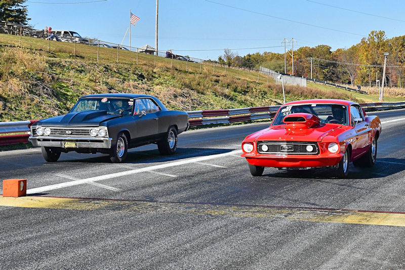 Two classic cars racing down a drag strip, one black and one red, with an American flag in the background.