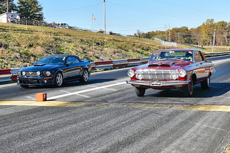A black modern sports car and a red classic car are racing down a drag strip, with a clear blue sky in the background.