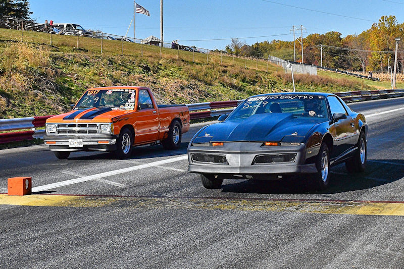 Two cars lined up at a drag racing track, one an orange pickup truck and the other a dark sports car, ready for a race.