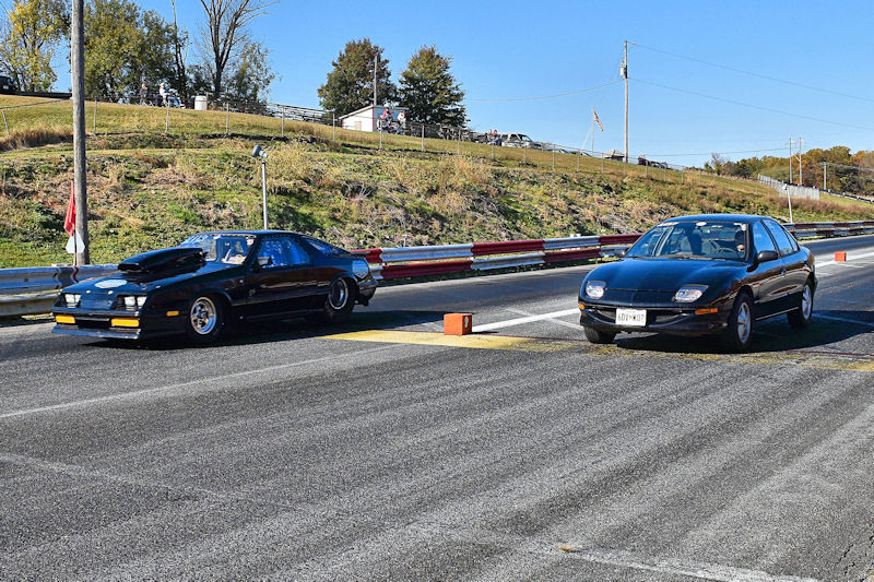 Two cars racing on a dragstrip, one with a raised hood and the other a compact sedan, under a clear blue sky.