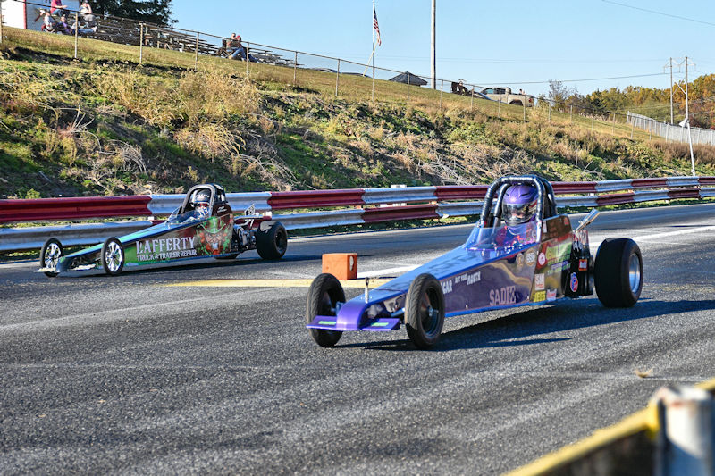 Two drag racing cars competing on a track on a sunny day, with one car in the foreground and the other in the background. The cars have distinct colorful designs, and the scene includes a grassy area and spectators in the background.
