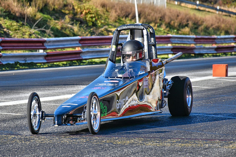 A black drag racing car with a transparent cockpit and a driver wearing a helmet, on a racetrack with barriers in the background.