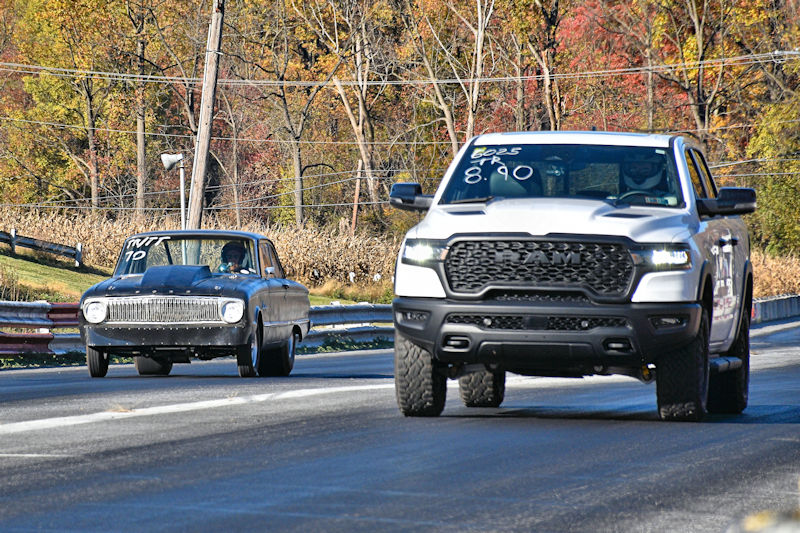 A classic black car racing alongside a modern white pickup truck on a racetrack, surrounded by autumn foliage.