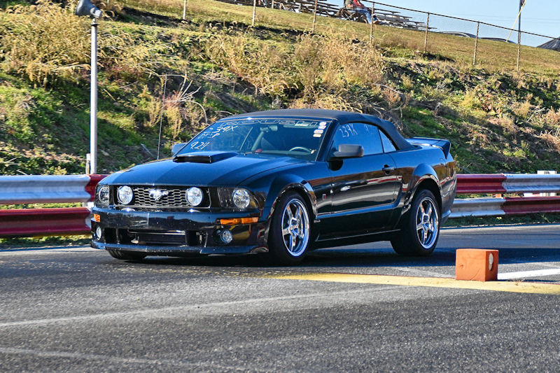 A black Ford Mustang convertible racing on a drag strip with a barrier and grassy area in the background.