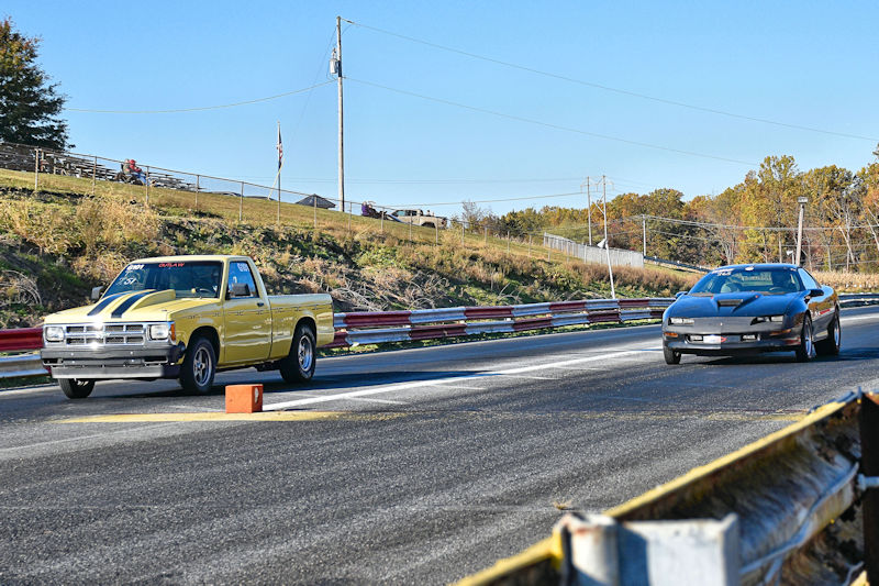 Two cars racing on a drag strip, with a yellow pickup truck on the left and a black sports car on the right.