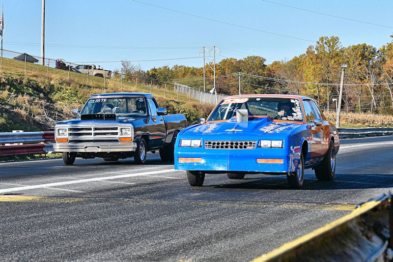 Two race cars competing on a drag strip, one is a black pickup truck and the other is a blue car with decals.