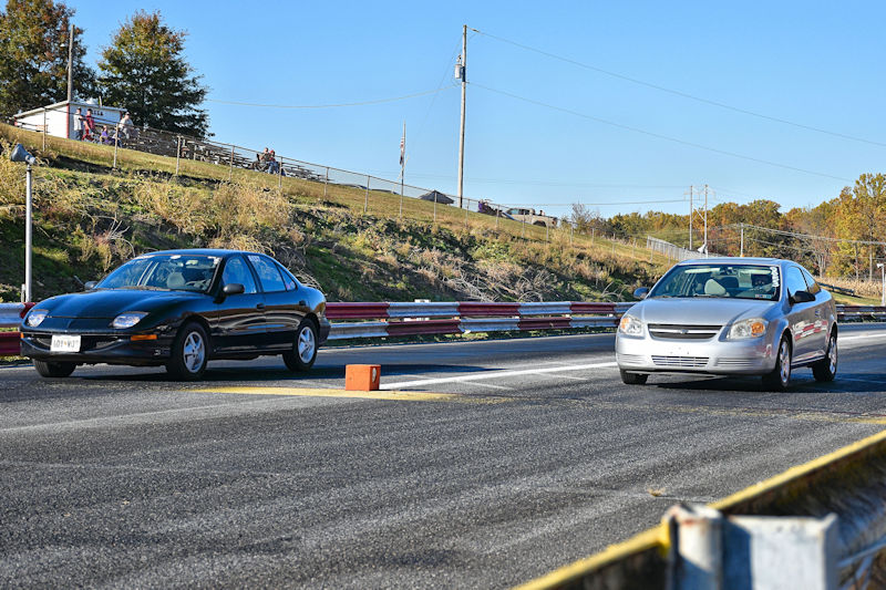 A black car and a silver car racing down an asphalt track with a starting light in the foreground.