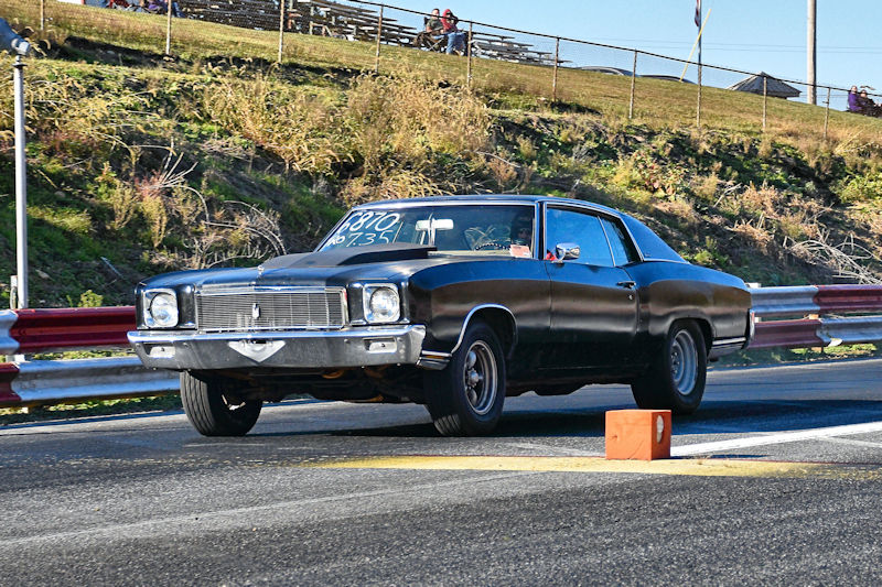 A black classic car racing down a drag strip with a starting line marker in the foreground.