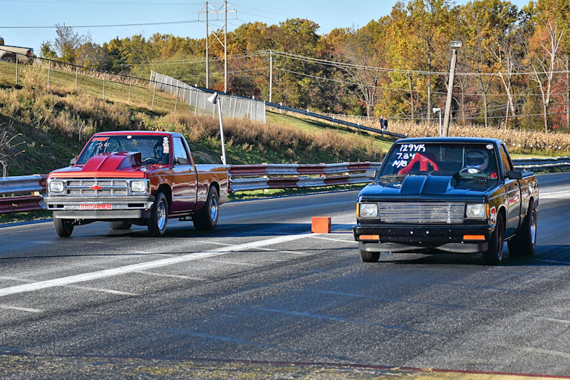 Two pickup trucks racing on a drag strip, with a tree-lined background and autumn foliage.