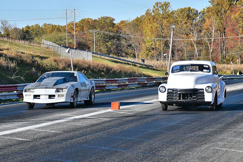 Two racing cars competing on a drag strip, with autumn foliage in the background. The left car is a modified coupe with a faded exterior, while the right car is a classic truck with a white paint job.