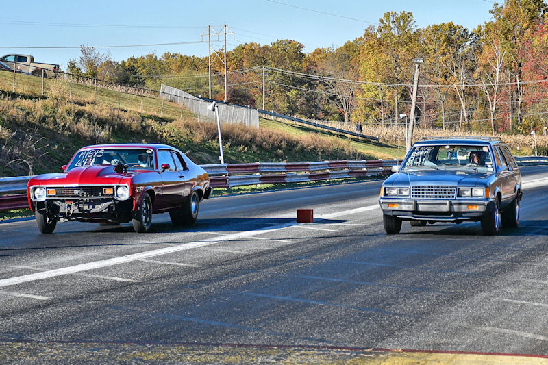 Two cars racing on a drag strip, with a red muscle car on the left and a gray car on the right.