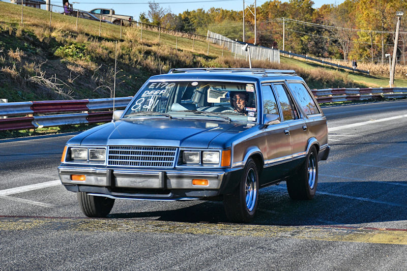A classic silver station wagon driving on a track with the driver smiling, surrounded by a green landscape and autumn trees in the background.