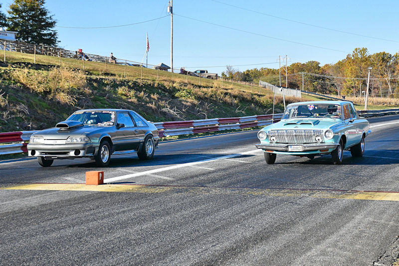 Two cars racing down a drag strip, one is a gray muscle car and the other is a light blue classic car.