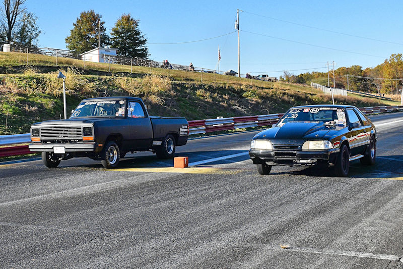 A black pickup truck and a black car lined up for a drag race on a racetrack, with the backdrop of grass and trees.