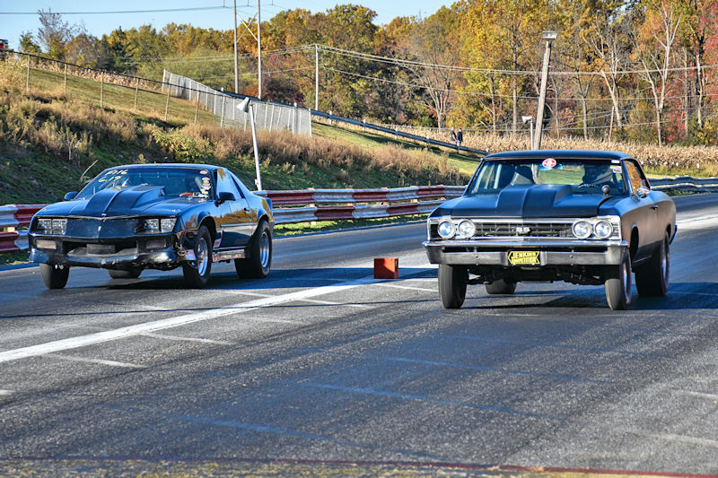 Two race cars lined up on a drag strip, preparing to race, with a tree and spectators in the background.