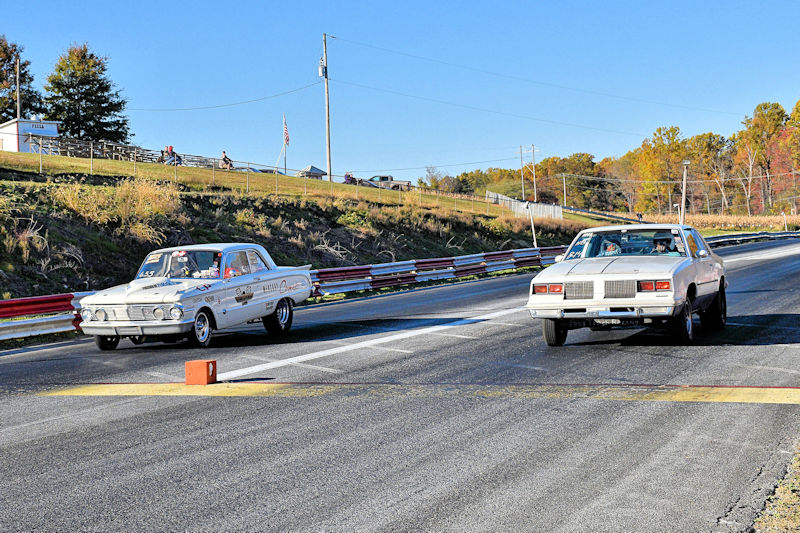 Two classic cars racing on a drag strip, with spectators in the background and autumn foliage along the sidelines.