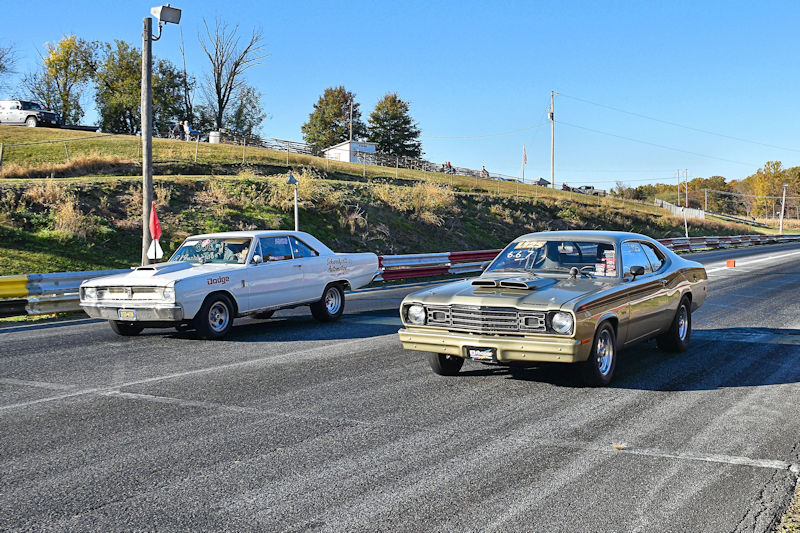 Two vintage cars lined up on a drag racing track, one in light gray and the other in olive green, under a clear blue sky.
