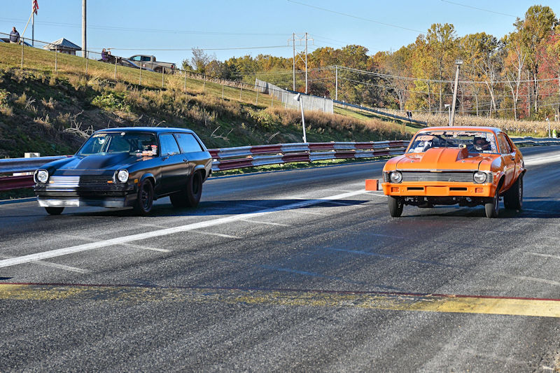 Two cars racing on a drag strip, one black and one orange, with spectators in the background and trees in fall colors.
