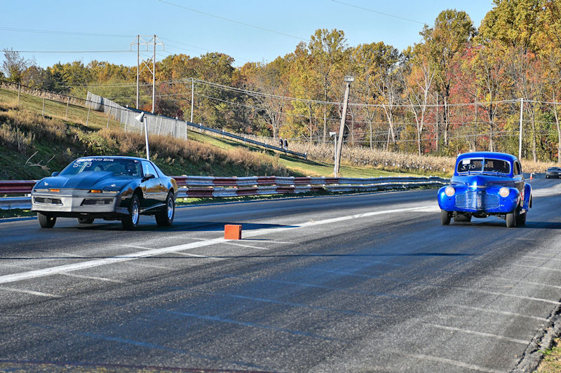 A black sports car and a blue vintage car racing on a drag strip with trees in the background.