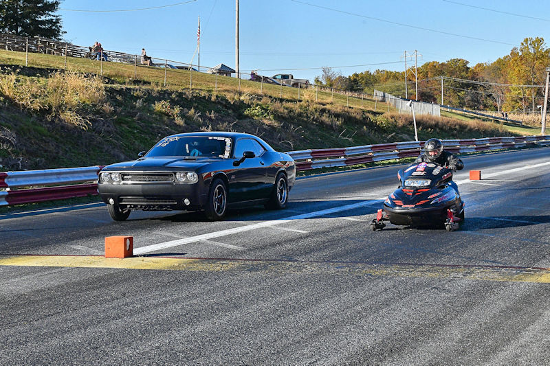 A black muscle car and a racing jet ski compete side by side on a drag racing track under a clear blue sky.