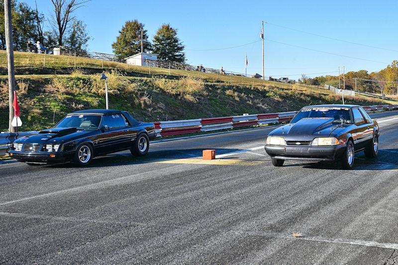Two cars racing on a drag strip, with a clear blue sky and spectators in the background.