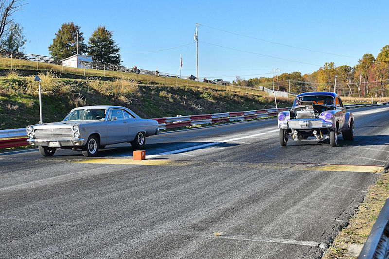 Two classic cars lined up for a drag race on a racetrack, with one silver car on the left and a purple car on the right.