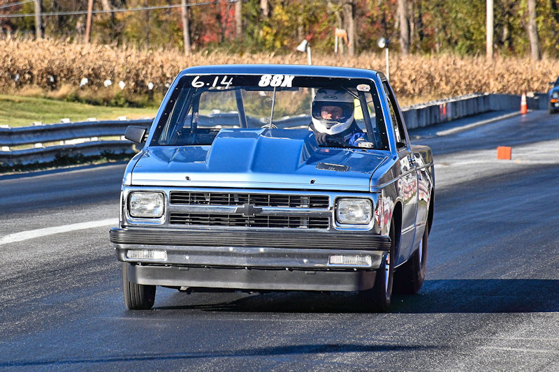 A racing truck with a blue finish racing down a track, with a driver wearing a helmet and racing gear. The truck displays the number 88X and has a time of 6.14 on its windshield.